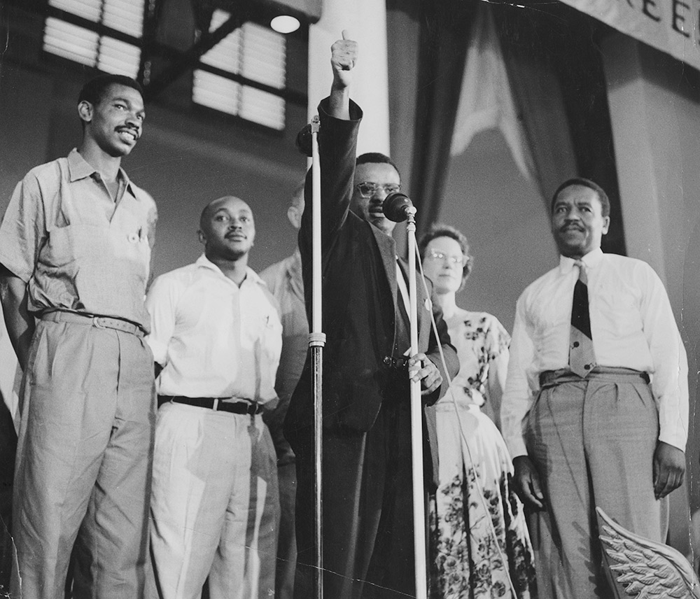 Mphahlele on stage at the All Africa People’s Conference, a Pan-Africanist Conference held in Accra, 1958. Also on stage are Alfred Hutchinson (far left), author of the significant exile memoir <em>Road to Ghana</em>, and Alfred Luthuli’s secretary, Mary-Louise Hooper (second from right). Mphahlele represented the ANC at the conference. This photo captures the moment at which the 200 delegates sang ‘Nkosi Sikelel’ iAfrika’, to which Mphahlele raises his thumb. Africa Media Online / Drum Photographer / Bailey’s African History Archive.